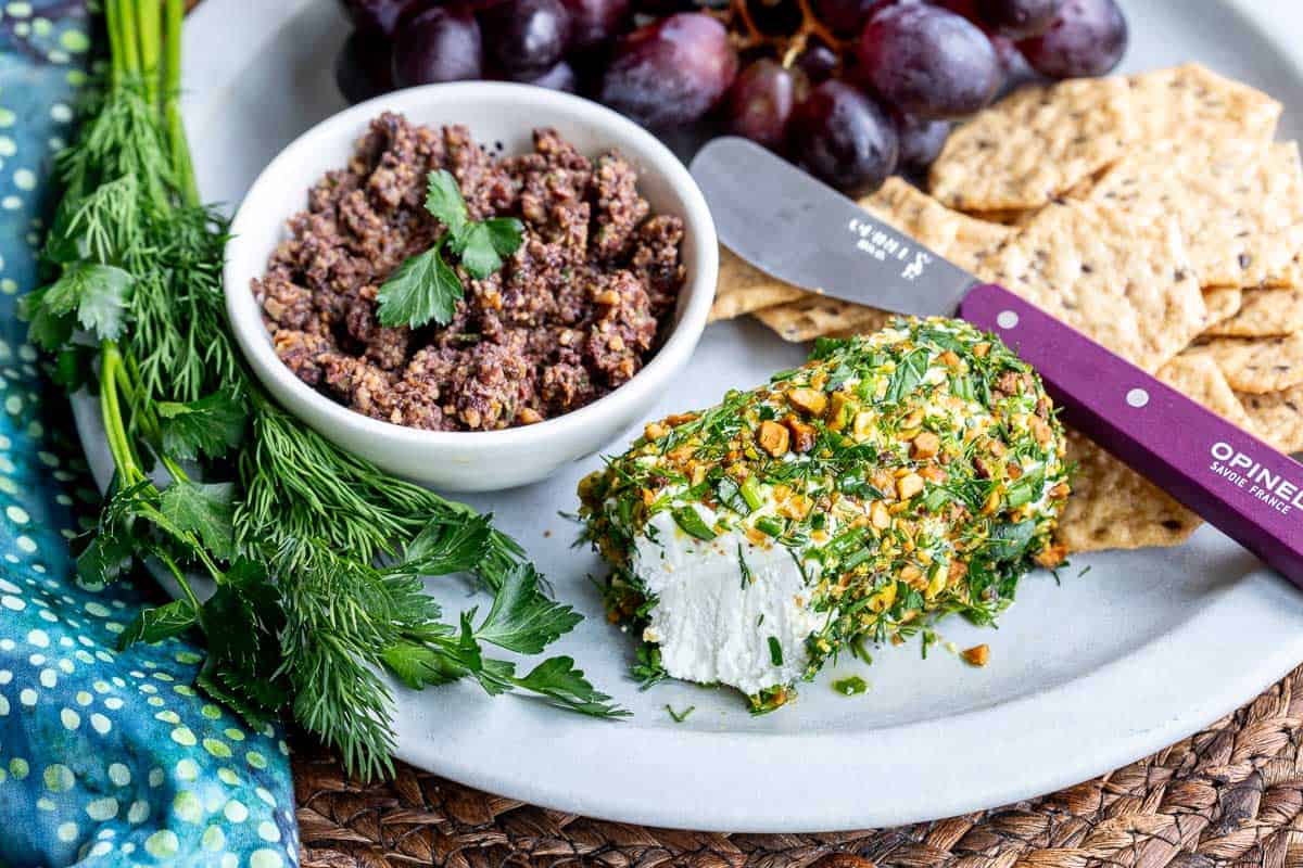 A plate with crackers, a log of herbed goat cheese coated in herbs and nuts, a bowl of tapenade, fresh dill, grapes, and a cheese knife.