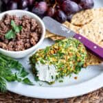 A plate with herbed goat cheese rolled in nuts and herbs, a bowl of olive tapenade, crackers, fresh herbs, and red grapes. A cheese knife rests on the plate.
