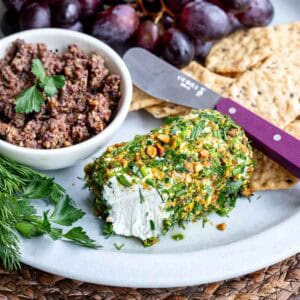A plate with herbed goat cheese rolled in nuts and herbs, a bowl of olive tapenade, crackers, fresh herbs, and red grapes. A cheese knife rests on the plate.