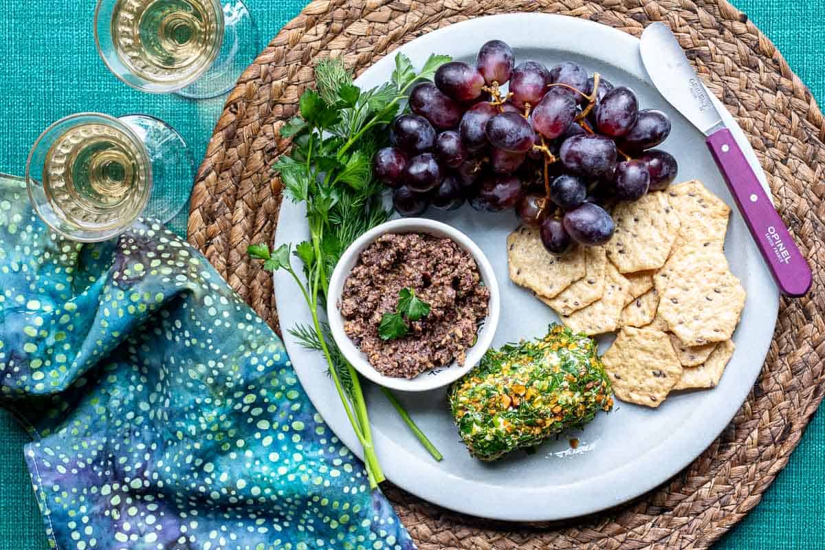 A plate with crackers, a herb-coated cheese log, a bowl of tapenade, red grapes, and fresh herbs, next to two glasses of white wine and a patterned napkin.