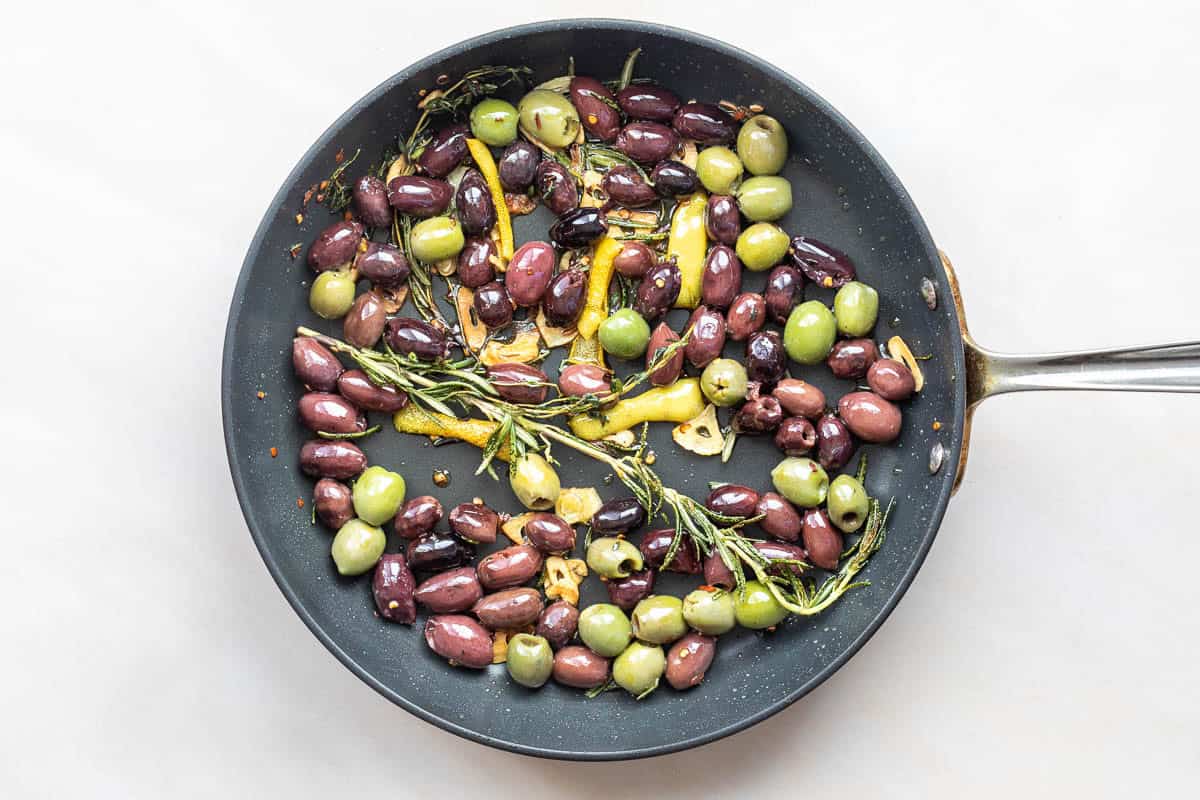 A frying pan with green and black olives, lemon slices, garlic, and rosemary sprigs on a white background.
