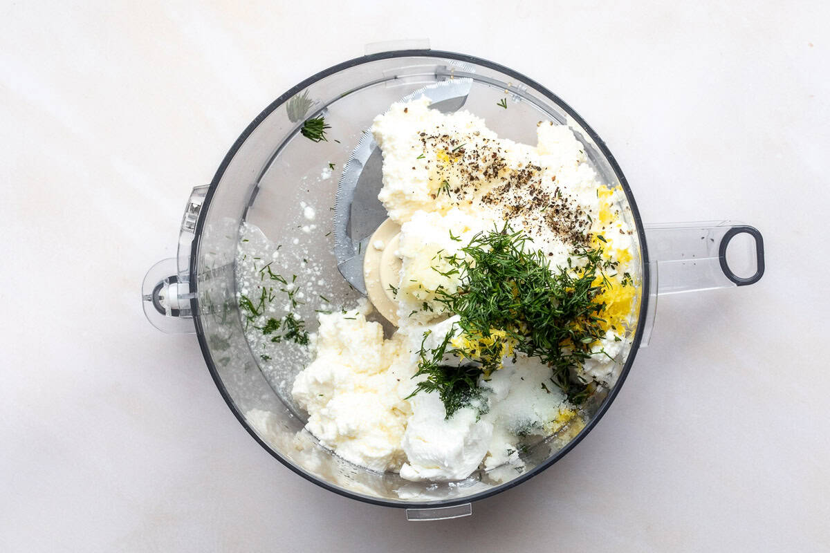 A food processor bowl with ricotta cheese, fresh dill, goat cheese, lemon zest, and black pepper, ready to be blended.