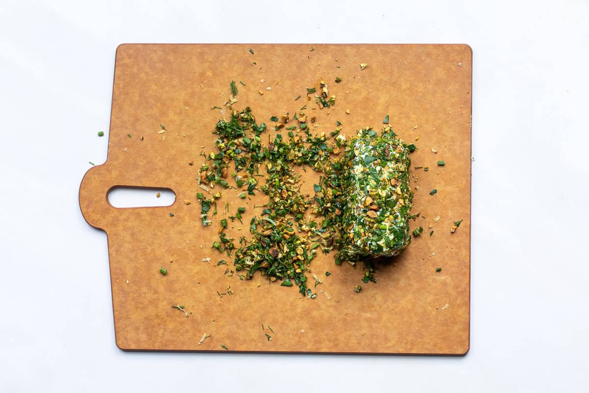 A log of herbed goat cheese with chopped herbs and pistachios scattered on a brown cutting board.