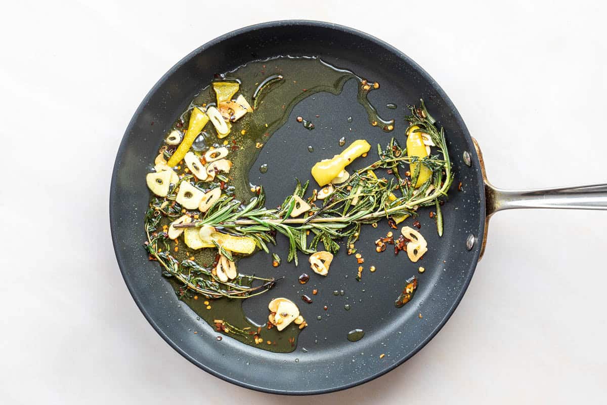 A black frying pan with olive oil, sliced garlic, lemon peel, red pepper flakes, and fresh rosemary on a white surface.