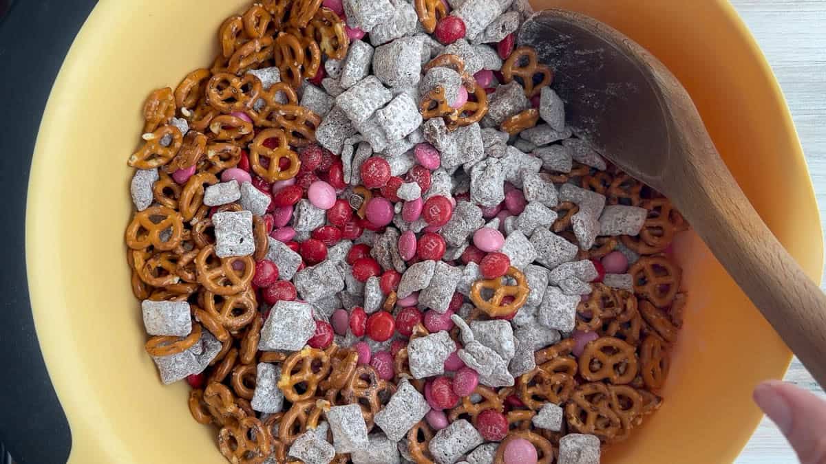 A mixing bowl containing pretzels, chocolate-covered candies, powdered sugar-coated cereal squares, and a wooden spoon.