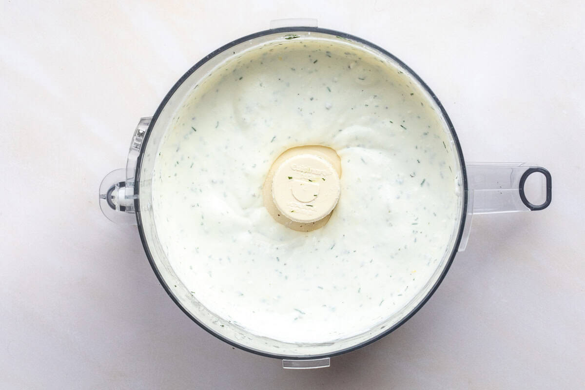 A food processor bowl filled with creamy whipped ricotta dip with green herb flecks sits on a light-colored surface.