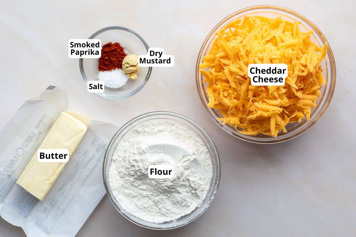 Five ingredients on a countertop: shredded cheddar cheese, flour, butter, smoked paprika, dry mustard, and salt, each in labeled bowls or containers.