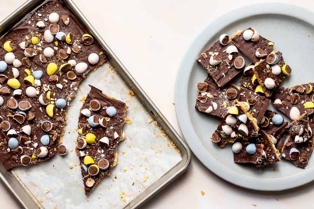 Saltine cracker toffee on baking sheet next to plate with broken pieces of saltine toffee.