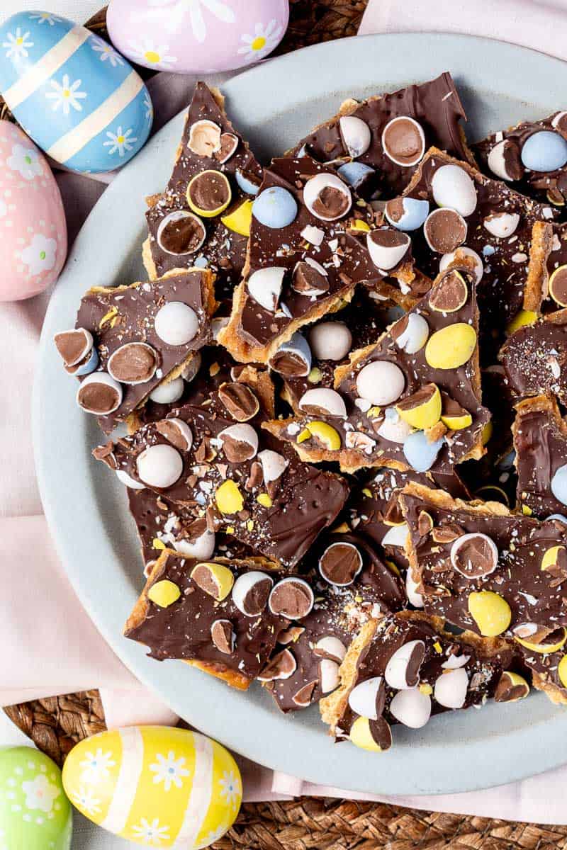 A plate of easter saltine cracker toffee with mini eggs, surrounded by decorative pastel Easter eggs.