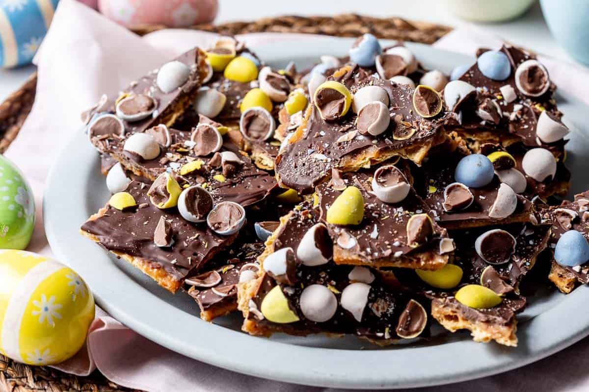 A plate of saltine cracker toffee bark topped with Cadbury mini eggs, set on a woven placemat with decorative Easter eggs nearby.
