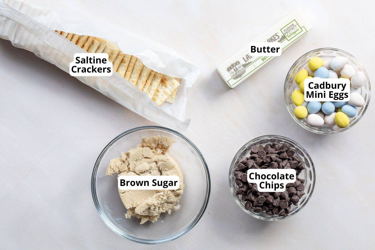 Saltine crackers, brown sugar, butter, chocolate chips, and Cadbury Mini Eggs are arranged in separate containers on a white surface.