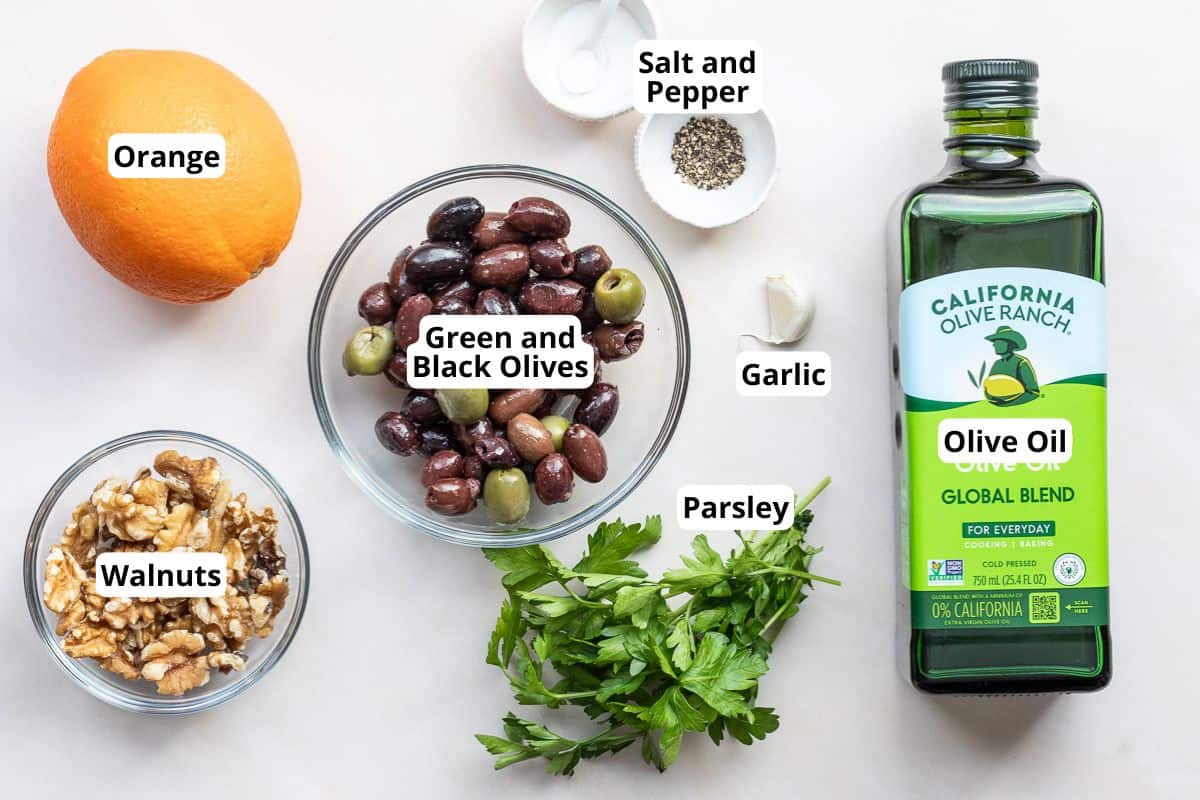 Ingredients including an orange, walnuts, green and black olives in a bowl, parsley, garlic, salt and pepper, and a bottle of olive oil, arranged on a light surface.