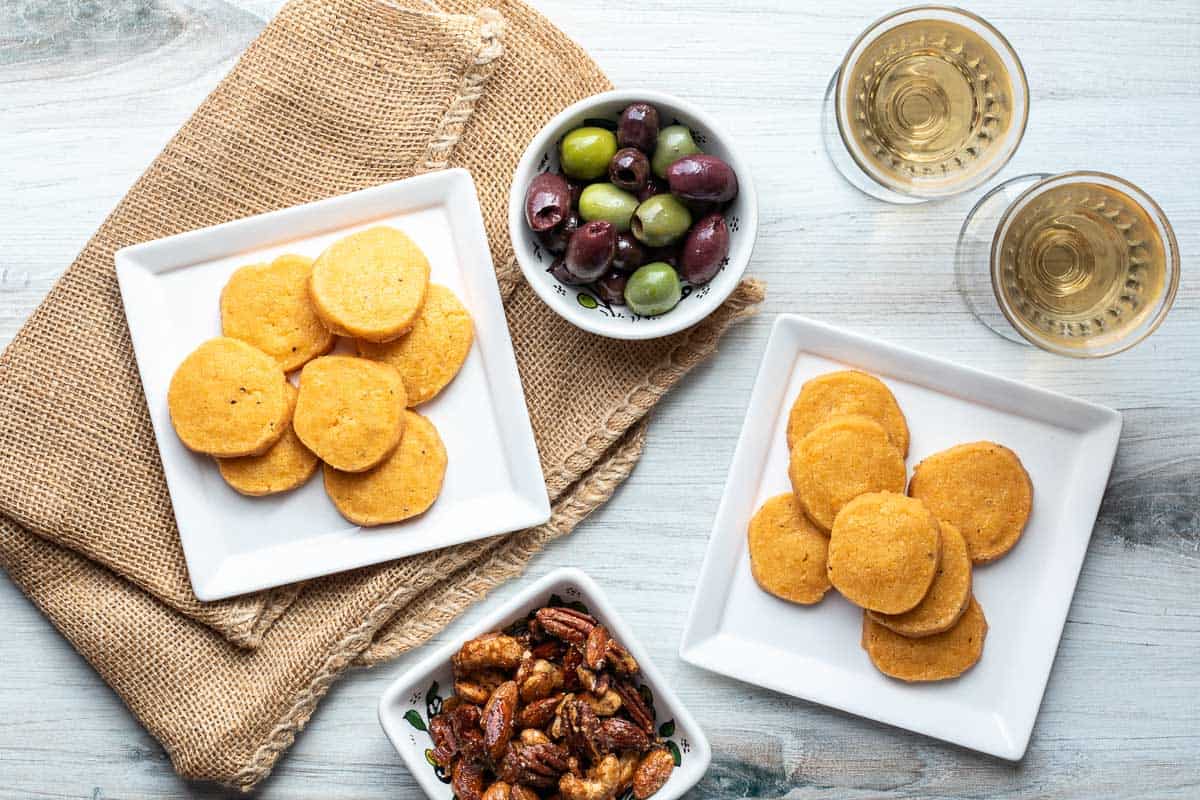 Two square plates of round homemade cheese crackers, a bowl of mixed olives, a bowl of candied nuts, and two glasses of white wine on a rustic surface with burlap cloth.