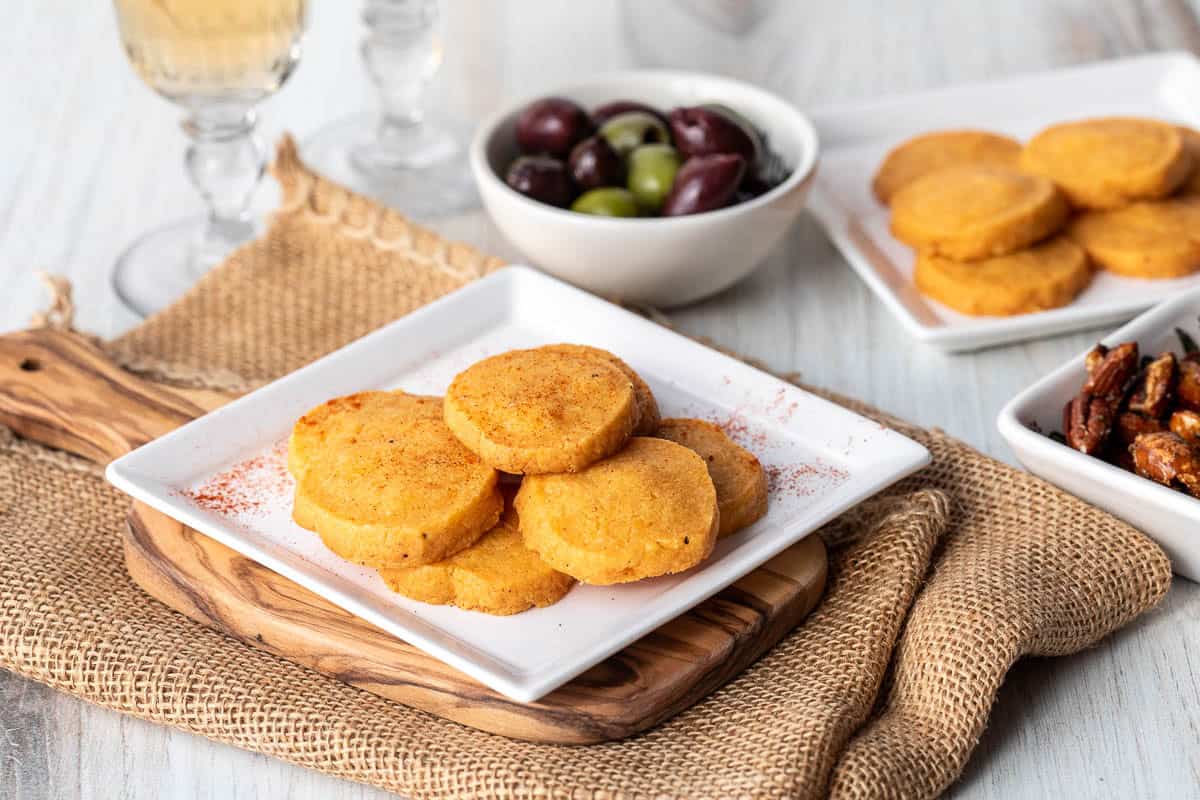 A plate of round cheese sablés is on a white square dish, with bowls of olives and other snacks and glasses of white wine in the background.
