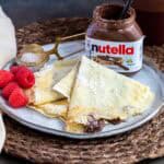 Plate with folded crepes filled with Nutella, raspberries on the side, a jar of Nutella, and a small sifter with powdered sugar.