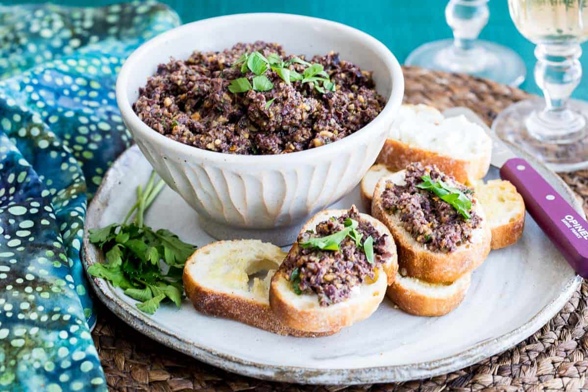 A bowl of olive tapenade garnished with herbs, surrounded by slices of baguette topped with tapenade, on a plate with a sprig of parsley.