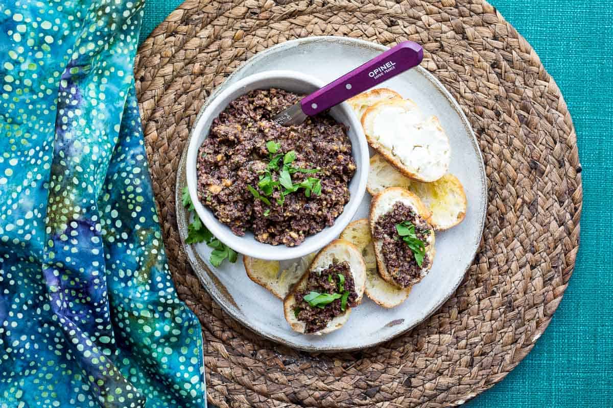 A bowl of olive tapenade with herbs and a knife sits on a plate with toasted bread slices, some topped with tapenade, on a woven placemat and blue patterned cloth.