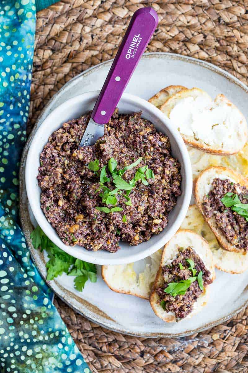 A bowl of olive tapenade with a purple-handled knife, served on a plate with toasted bread slices, some topped with tapenade and fresh herbs.