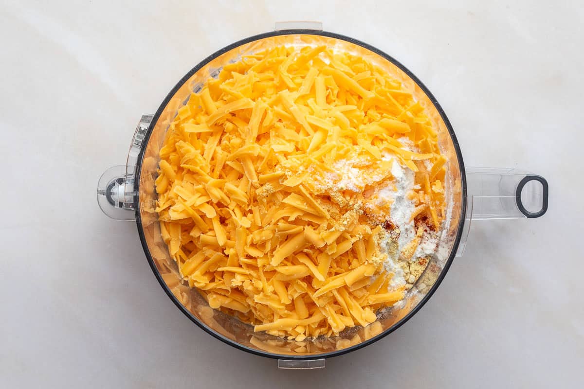 Food processor bowl filled with grated cheddar cheese, flour, and seasonings, viewed from above on a light surface.