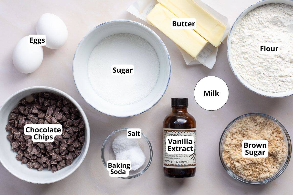 Baking ingredients arranged on a counter: eggs, sugar, butter, flour, chocolate chips, salt, baking soda, vanilla extract, milk, and brown sugar, each labeled.