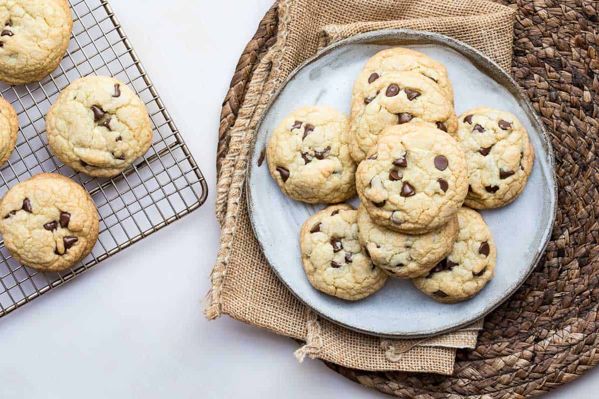 A plate with several brown butter chocolate chip cookies on a woven mat, next to a cooling rack with more cookies on a white surface.