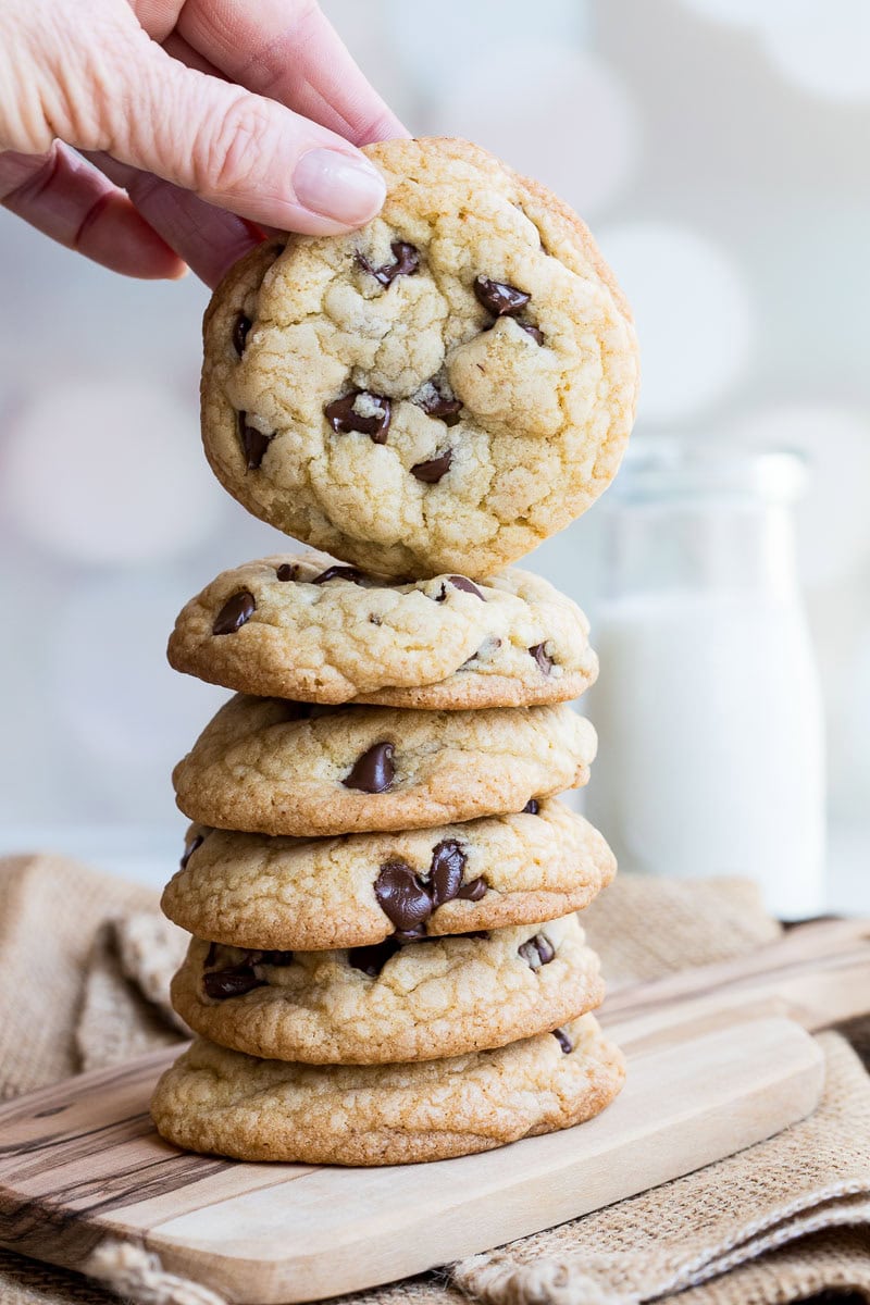A hand holds a brown butter chocolate chip cookie above a stack of five cookies on a wooden board, with a glass bottle of milk in the background.
