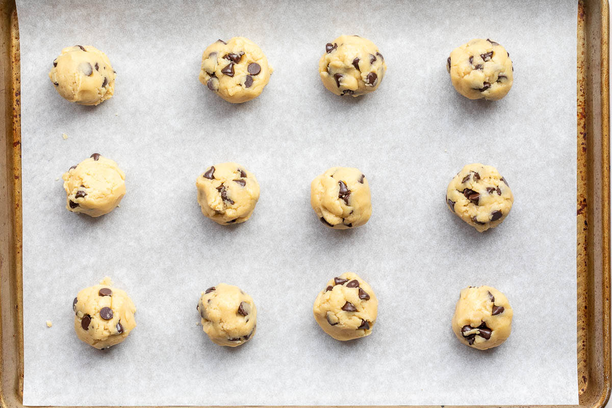 Twelve balls of brown butter chocolate chip cookie dough are evenly spaced on a parchment-lined baking sheet, ready to be baked.