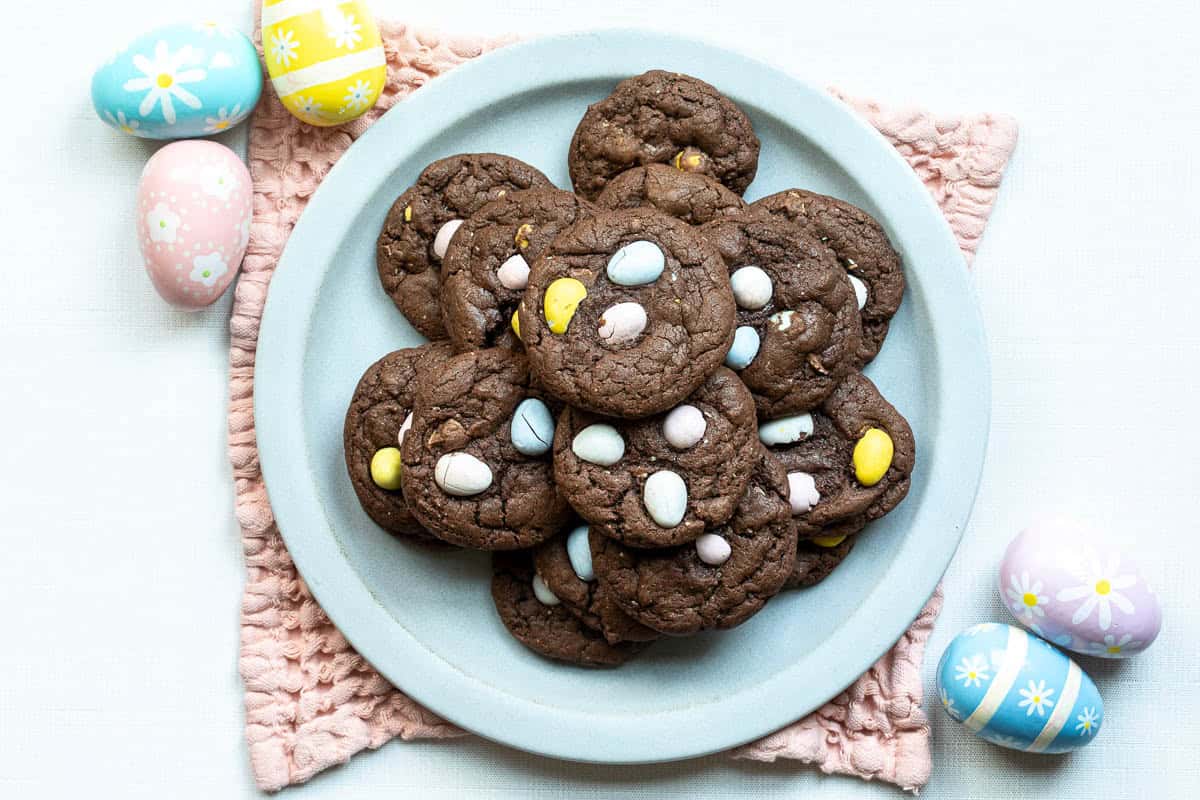 A plate of chocolate cookies topped with Cadbury mini eggs sits on a pink cloth, surrounded by decorated Easter eggs.