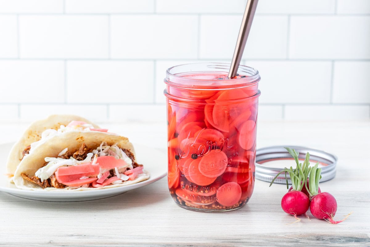 A glass jar filled with sliced pickled radishes in pink brine, with a spoon inside. The jar sits on a white surface next to a radish and a partially visible sandwich.