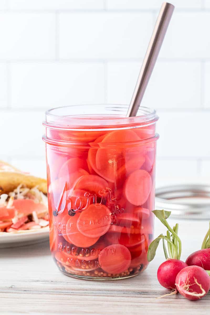 A glass jar filled with sliced pickled radishes in pink brine, with a metal spoon inside. Fresh radishes and a plate of food are in the background.