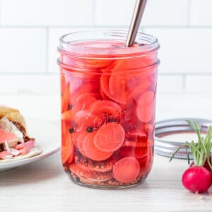 A jar of pickled radishes with a fork inside sits next to fresh radishes and a plate with tacos topped with pickled radishes.