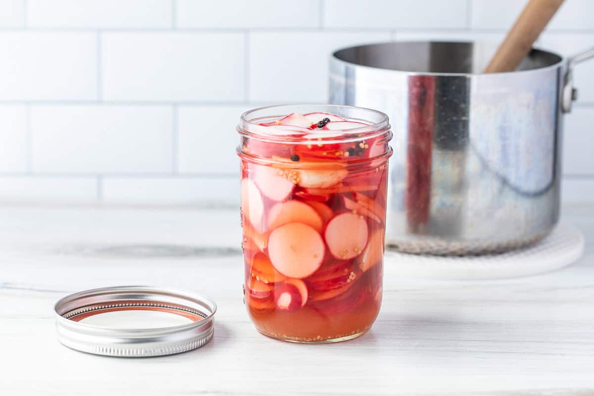 A glass jar filled with sliced radishes in pickling brine sits on a white surface next to its lid, with a metal pot and wooden spoon in the background.