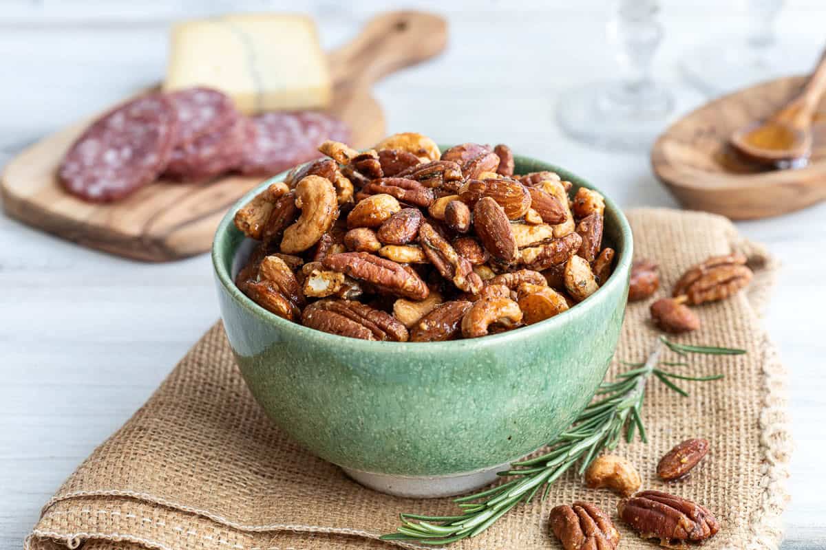 A green bowl filled with mixed honey roasted nuts sits on a burlap mat, with rosemary, sliced salami, and cheese on a wooden board in the background.