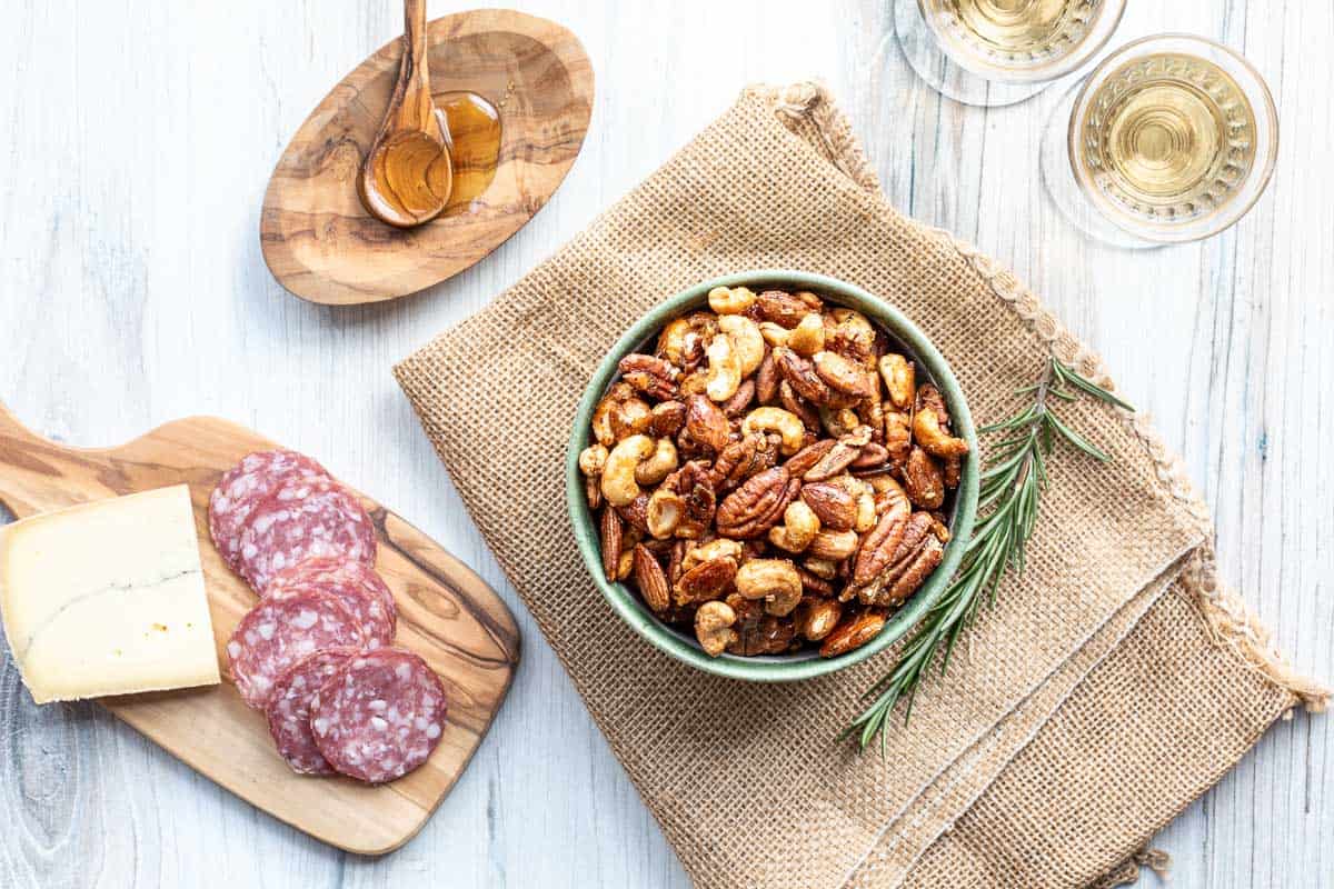 A bowl of mixed roasted nuts, sliced salami, a wedge of cheese on a wooden board, two glasses of white wine, and a dish of honey on a burlap cloth.