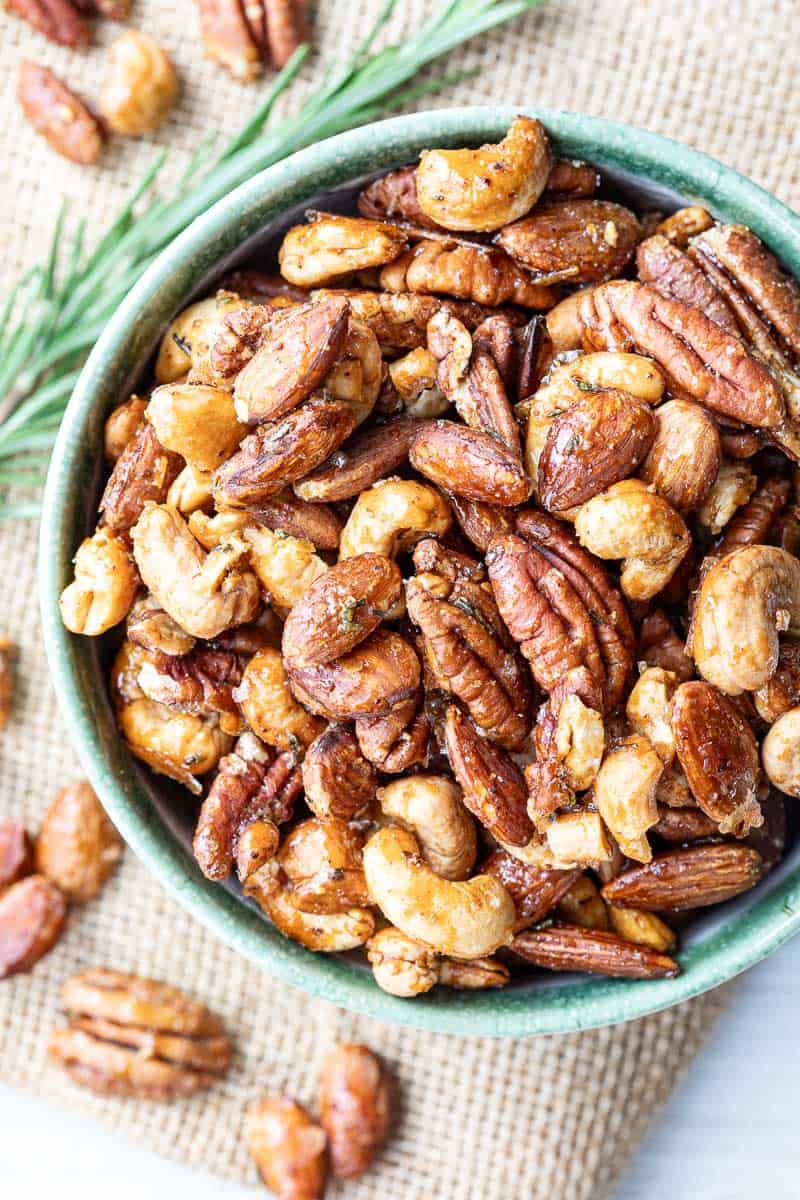 A bowl filled with a mixed assortment of roasted nuts, including pecans, cashews, and almonds, on a burlap surface with sprig of rosemary.