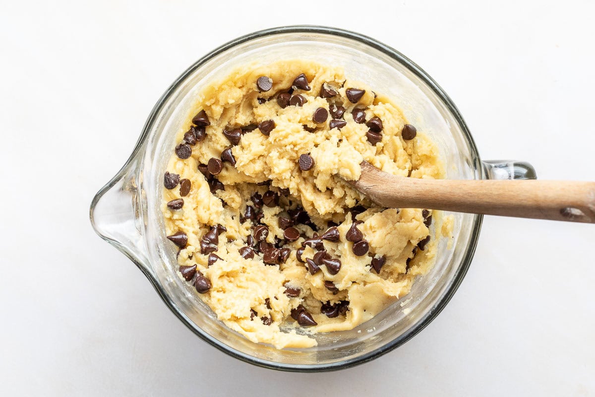 A glass bowl filled with brown butter chocolate chip cookie dough being mixed with a wooden spoon on a white surface.