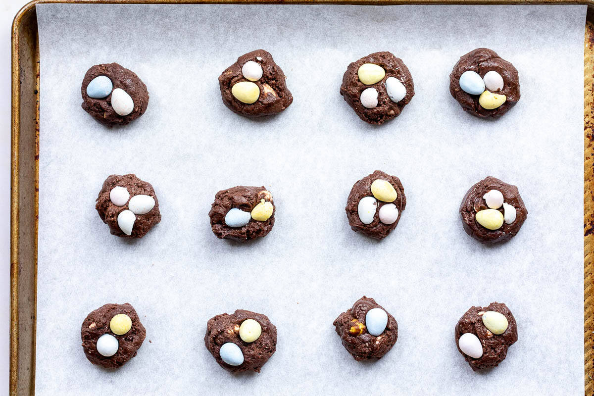 Fifteen unbaked chocolate cookies topped with Cadbury mini eggs are arranged in rows on a parchment-lined baking sheet.