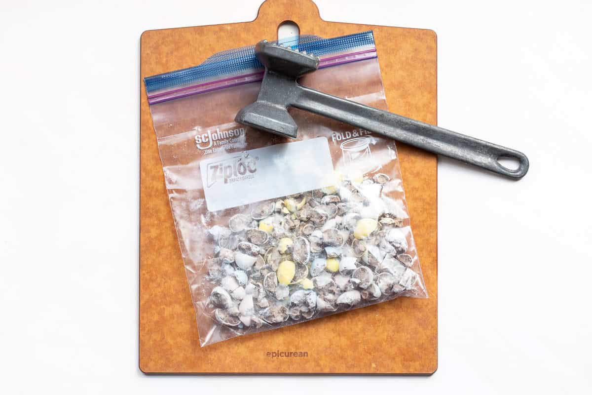 A sealed plastic bag containing crushed Cadbury mini eggs is placed on a cutting board with a metal meat mallet resting on top.