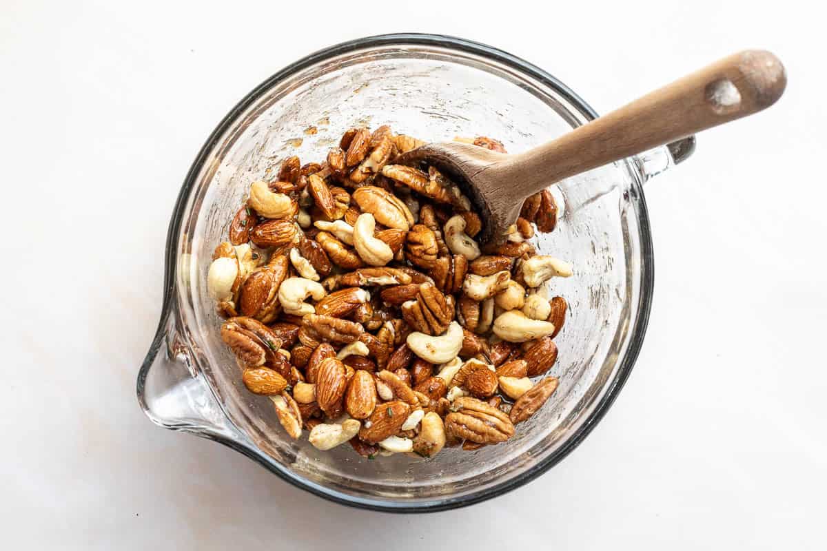 A glass mixing bowl filled with assorted nuts, including almonds, cashews, and pecans, being stirred with spices and sugar and herbs with a wooden spoon.