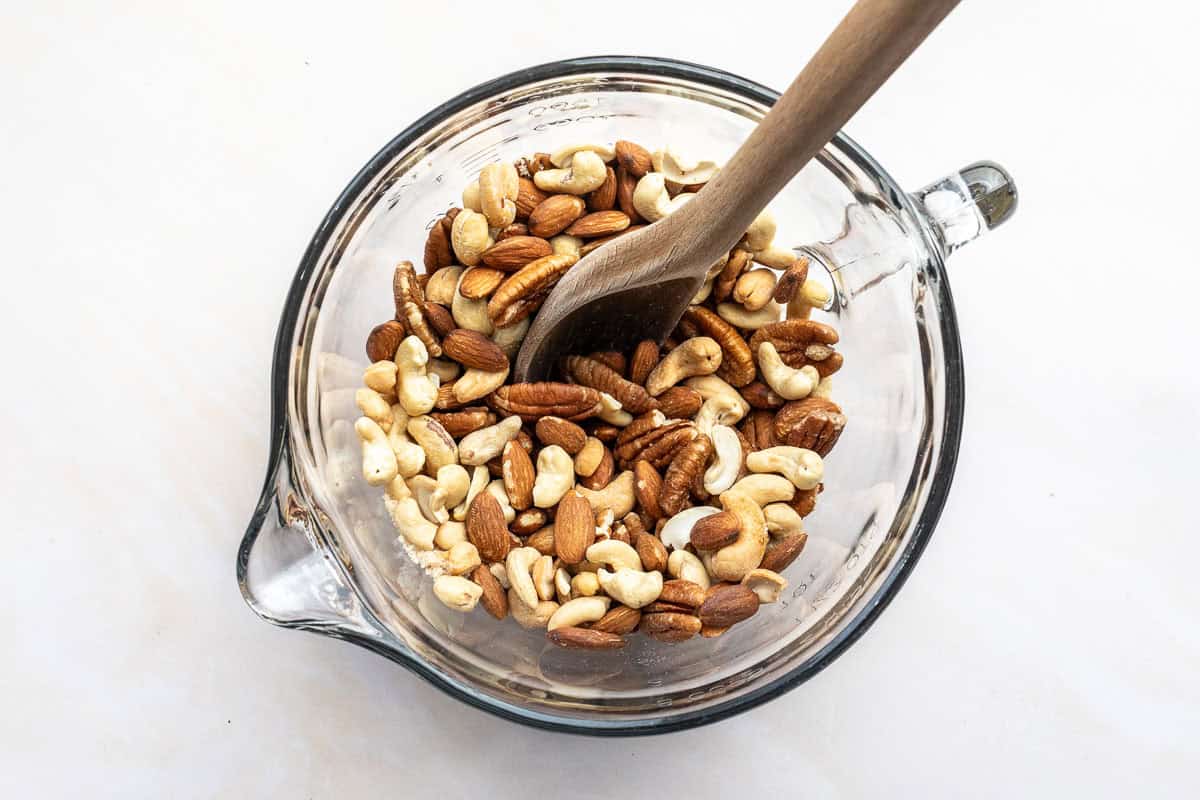 A glass bowl filled with a mixture of raw nuts, including cashews and almonds, being stirred with a wooden spoon.
