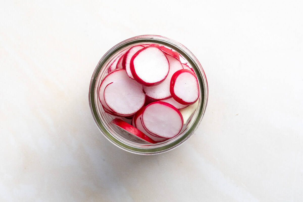A glass jar filled with sliced red radishes and garlic, viewed from above on a light-colored surface.