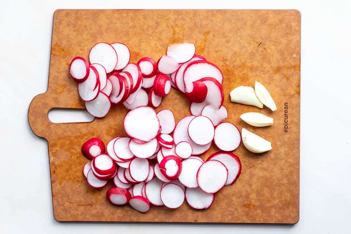 Sliced radishes and peeled garlic cloves arranged on a wooden cutting board.