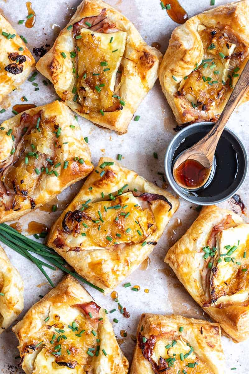 Golden ham and cheese puff pastries topped with caramelized onions and chopped chives are arranged on parchment paper next to a small bowl of sauce with a wooden spoon.