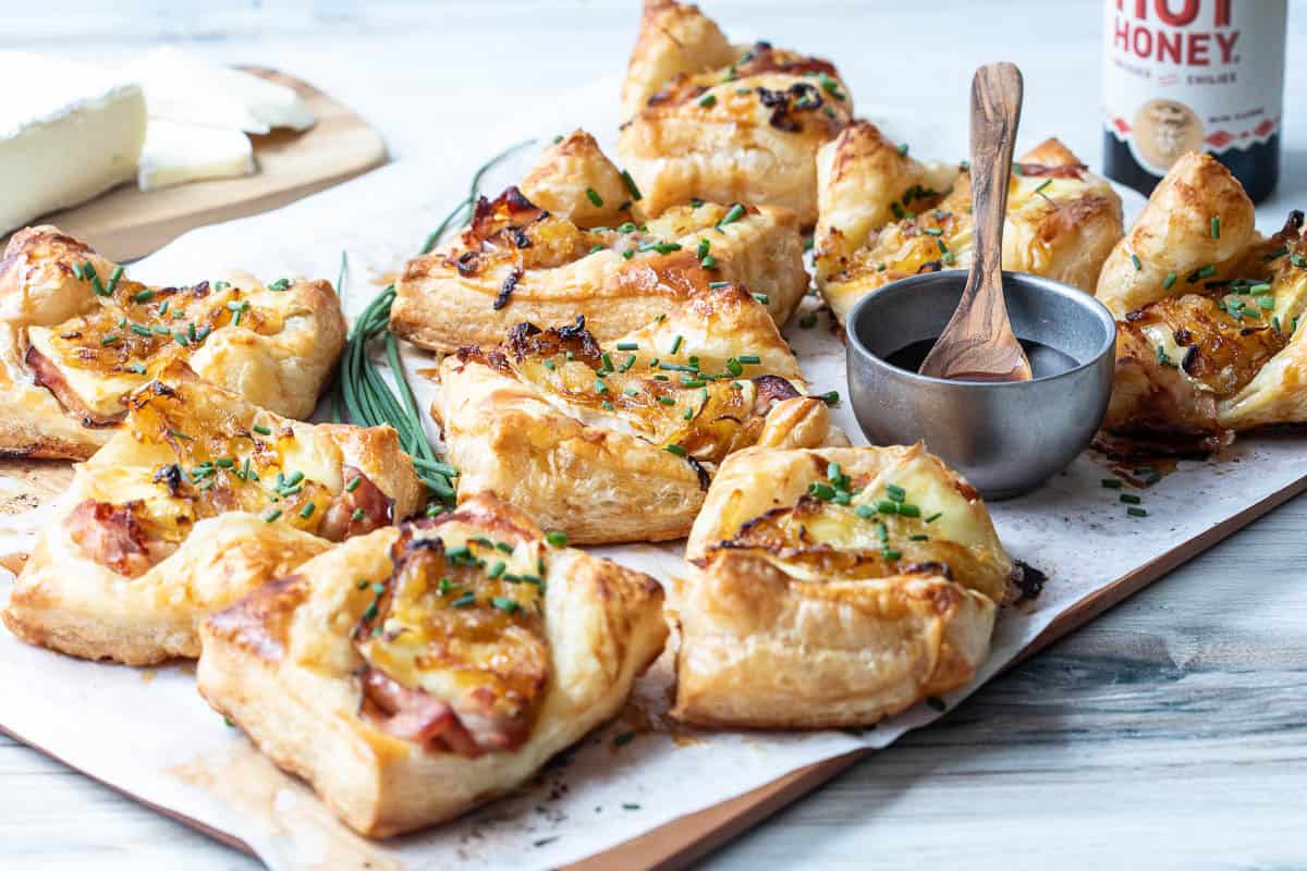 A wooden board with several puff pastry tarts topped with caramelized onions, ham, cheese, herbs, and a hot honey dip in a small metal bowl, with cheese slices in the background.