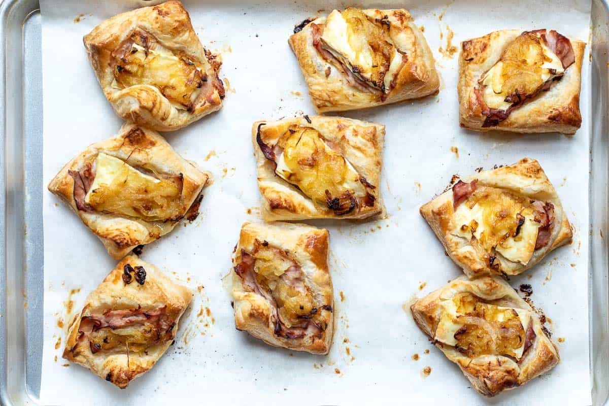 Nine ham and cheese in puff pastry parcels topped with caramelized onions on parchment paper atop a baking sheet.