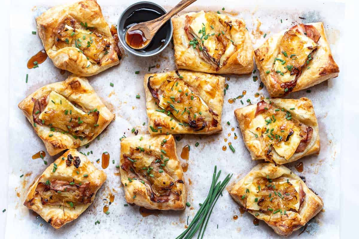 Nine puff pastry squares topped with melted cheese, ham, and herbs are arranged on parchment paper, with a small bowl of sauce and a wooden spoon nearby.