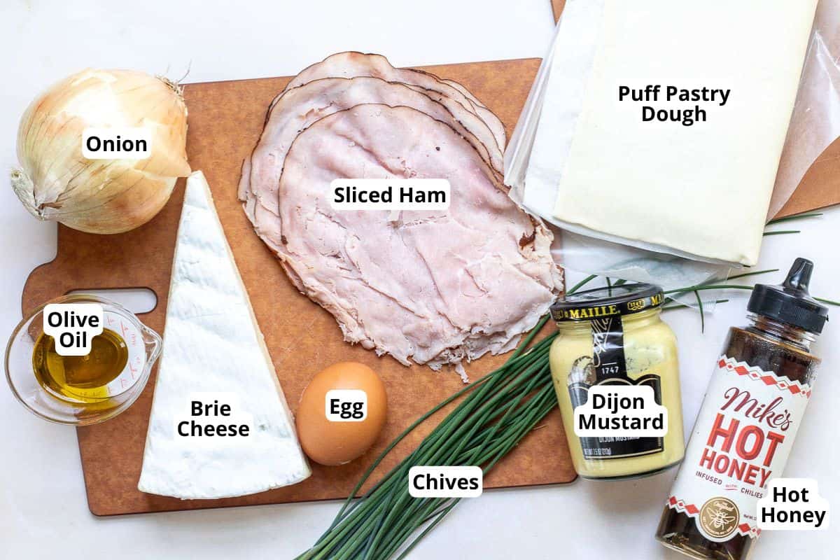 Ingredients for Ham and Cheese Puff Pastries are arranged on a cutting board, including onion, olive oil, brie cheese, sliced ham, egg, chives, puff pastry dough, Dijon mustard, and hot honey.
