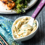 A small white bowl filled with miso butter with chives and a purple-handled spreading knife, placed on a wooden table near a blue patterned napkin.