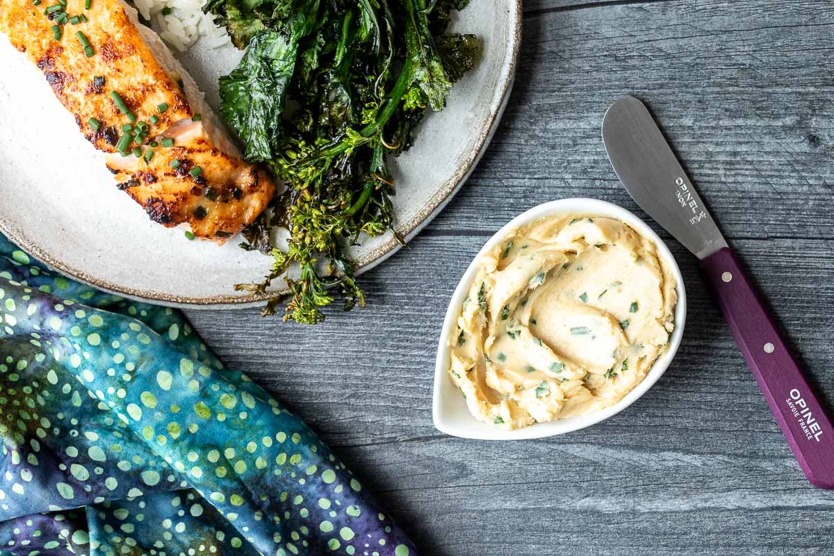 Plate with grilled salmon, crispy greens, and rice next to a dish of herb miso butter and a butter knife on a gray wooden table with a blue patterned cloth.