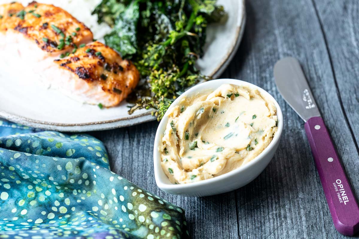 A bowl of herb miso butter sits next to a plate with cooked salmon and greens on a wooden table, with a butter knife and patterned cloth nearby.